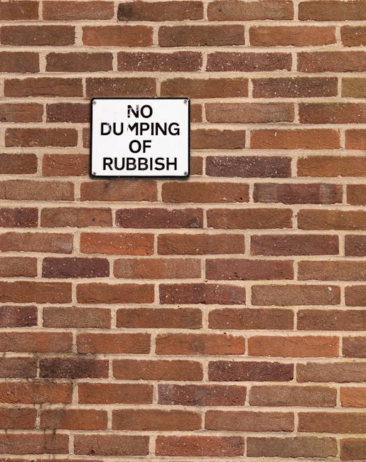 Close-up view of a white rectangular metal sign mounted on a red brick wall, with black text that reads 'NO DUMPING OF RUBBISH'. The brick wall features a uniform pattern of reddish-brown bricks with darker mortar joints, and the sign is positioned towards the upper left section of the image. The lighting appears natural, casting subtle shadows on the wall, emphasizing the textured surface of the bricks and the sign’s slightly reflective surface. This scene relates to an environment where waste management or rubbish removal is a concern, often addressed through private clearance services such as those offered by Rubbish Removal Harringay, which might be employed for on-site clearance and preventing illegal dumping in residential or commercial areas.
