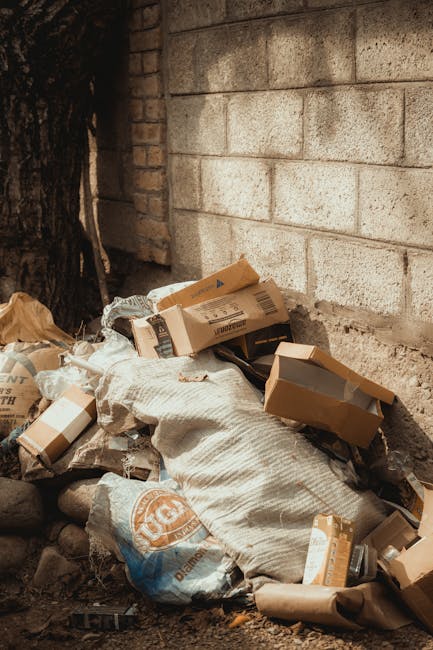 A pile of discarded cardboard boxes, some flattened and others partially unassembled, piled against a weathered brick wall outdoors. The boxes vary in size and are made of brown corrugated cardboard, with visible printed labels and barcodes. Several of the boxes are open or torn, with some containing shredded or crumpled packaging material. The ground beneath the pile is uneven, composed of dirt and small rocks, and there is a large tree trunk nearby on the left side of the image. The background includes a light-colored brick wall with a rough texture, and part of a dark shadow cast by the tree. The scene appears to depict an area prepared for waste disposal or collection, aligning with independent rubbish removal or on-site clearance activities, as part of private waste handling services offered by Rubbish Removal Harringay.