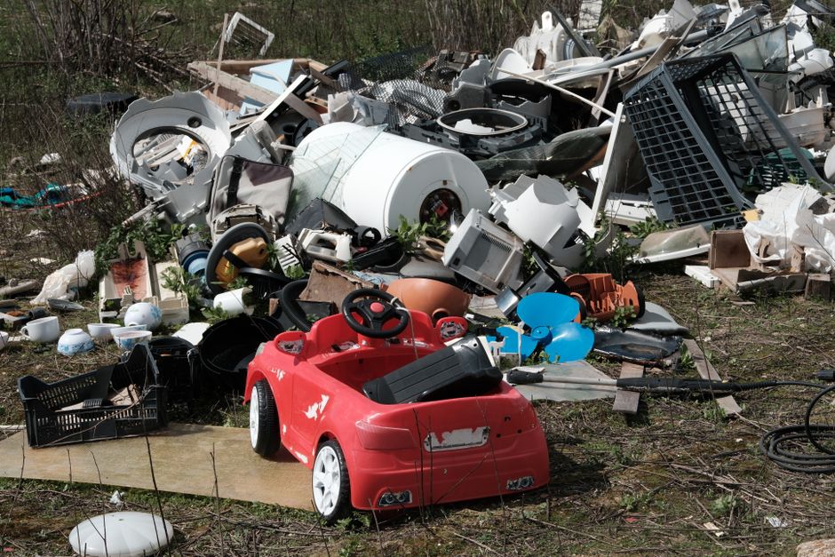 A collection of discarded household and electronic waste scattered across an outdoor area, with a focus on a red plastic children's ride-on toy positioned at the front center. The toy, designed to resemble a small vehicle with a black seat, steering wheel, and white wheels, shows signs of wear including scratches and chipped paint. Surrounding the toy are various broken appliances, such as a white washing machine drum, a black plastic crate, and a mixture of items including ceramic dishes, plastic containers, and metal parts. The background features a jumble of scrap materials, including metal fencing, plastic piping, and disassembled household items, all resting on uneven ground covered with patches of grass, weeds, and dirt. The scene is outdoors, likely in a rural or semi-urban setting, illuminated by natural daylight, emphasizing the disorderly removal of the waste. The cluttered arrangement and presence of household debris reflect an on-site clearance or private waste collection process typical of rubbish removal services, such as those offered by Rubbish Removal Harringay.