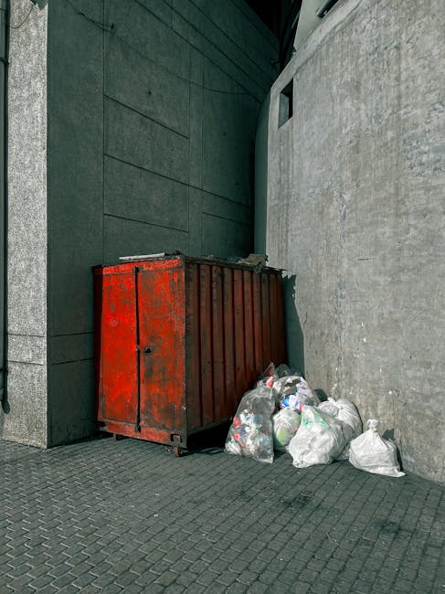A weathered red metal skip with visible rust and scratches is positioned against a dark green wall on the left and a light grey concrete wall on the right in an urban outdoor setting. The skip is rectangular with a closed lid on top, and it rests on a paved surface composed of small dark grey bricks. Several white plastic rubbish bags, some slightly flattened and others bulging, are stacked and piled next to the skip, spilling onto the ground in front of it. The bags appear to be filled with mixed waste, possibly household and general rubbish. The scene is illuminated by natural light, casting minimal shadows, suggesting an overcast sky. The setting seems to be a back alley or side street area, with clean, flat surfaces and minimal environmental features visible, reflecting a typical private waste disposal site suitable for independent rubbish removal services, such as those offered by Rubbish Removal Harringay.