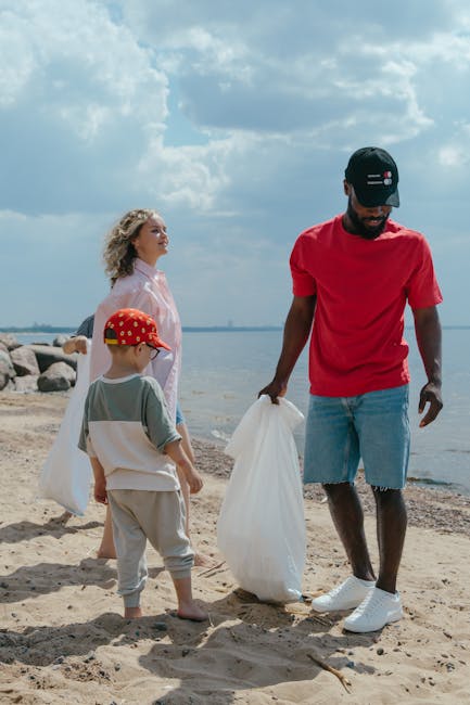 A group of three individuals, including a man and two children, are engaged in a litter-picking activity on a sandy beach near a body of water. The man, positioned on the right, is wearing a red T-shirt, light blue distressed denim shorts, white sneakers, and a black cap with red, white, and blue stripes; he is holding a large white plastic rubbish bag in his right hand, actively collecting debris from the sand. The two children stand on the left; the girl, with curly blonde hair, is dressed in a light pink shirt, and the boy is wearing a grey T-shirt, beige pants, a red baseball cap with white polka dots, and is holding a waste bag in his left hand. In the background, there are large rocks along the shoreline, and the sky above is partly cloudy with bright sunlight illuminating the scene. The environment suggests an initiative to remove litter from the beach, aligning with private waste collection efforts such as those by Rubbish Removal Harringay, contributing to cleaner outdoor spaces.