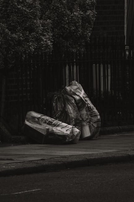 Two large black plastic rubbish bags, filled and tied at the top, sit on the pavement beside a black metal fence and a sidewalk curb, with a background of dense leafy trees and the dark silhouette of a building or structure behind the fence. The bags appear to contain household waste or discarded materials, with some visible branding or writing on their surfaces. The scene is captured in natural daylight, with the bags positioned in an outdoor urban environment, indicative of private waste disposal or alternative rubbish removal methods that Rubbish Removal Harringay may facilitate. The overall setting suggests an area awaiting collection, with the bags placed on the street for collection or disposal, reflecting typical small-scale rubbish clearance practices outside residential properties.