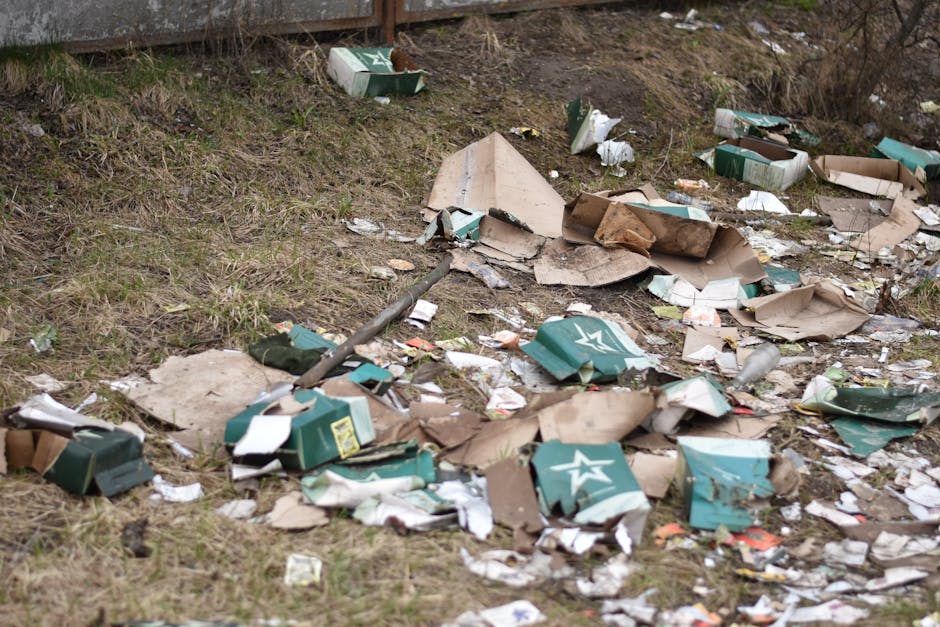 A patch of ground near a grassy area and a wooden fence is visibly littered with scattered cardboard boxes, some torn open and crumpled, along with various paper and plastic packaging. The cardboard boxes are primarily brown and green, with some displaying printed logos and labels; they vary in size and appearance, with some flattened and others partially intact. Among the debris, there are small empty plastic bottles and other miscellaneous waste items. The ground is uneven, with patches of grass and dirt, and the litter appears to have been recently discarded or blown into this outdoor space. The scene reflects an instance of improper waste disposal, suggesting the need for professional rubbish removal services such as those offered by Rubbish Removal Harringay, to effectively clear the accumulated waste from this area and maintain cleanliness and environmental standards.