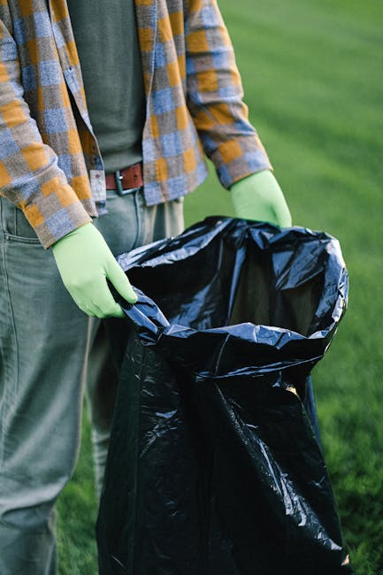 Two large black plastic rubbish bags, filled and tied at the top, sit on the pavement beside a black metal fence and a sidewalk curb, with a background of dense leafy trees and the dark silhouette of a building or structure behind the fence. The bags appear to contain household waste or discarded materials, with some visible branding or writing on their surfaces. The scene is captured in natural daylight, with the bags positioned in an outdoor urban environment, indicative of private waste disposal or alternative rubbish removal methods that Rubbish Removal Harringay may facilitate. The overall setting suggests an area awaiting collection, with the bags placed on the street for collection or disposal, reflecting typical small-scale rubbish clearance practices outside residential properties.