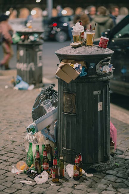 A black, cylindrical municipal rubbish bin with a domed lid is positioned on a cobblestone street, surrounded by scattered rubbish. On top of the bin, there are various discarded items, including used paper cups with plastic lids, some with leftover drinks, and a crumpled paper napkin. The front of the bin has a small door for waste disposal, and the surface shows weathered textures. Around the base of the bin, multiple glass bottles, some with labels in red and green, are tipped over or left amidst crumpled tissues and other debris. Behind the bin, a large grey trash bag leans against it, filled with additional waste. The background features a blurred urban scene with pedestrians, parked cars, and a green public trash bin, indicating an outdoor city environment. The overall scene depicts a typical street-side rubbish accumulation, highlighting the importance of efficient waste removal services, as managed by Rubbish Removal Harringay, to maintain public cleanliness and support alternative waste management methods in urban areas.