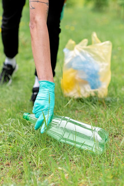 A person wearing turquoise gloves is actively picking up a clear plastic bottle from a grassy outdoor area, with their arm and hand visible in the foreground. In the background, there is a yellow plastic rubbish bag filled with various litter, including blue items, resting on the grass. The scene appears to be part of a cleanup activity in a park or similar green space, with lush grass and some yellow flowers visible around. The individual involved is wearing dark clothing, and the overall environment is well-lit, indicating daytime. This visual context relates to environmentally conscious waste collection, including private rubbish removal services like those provided by Rubbish Removal Harringay, that focus on independent disposal and on-site clearance of recyclable materials and general waste in outdoor settings.