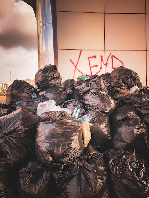 A black, cylindrical municipal rubbish bin with a domed lid is positioned on a cobblestone street, surrounded by scattered rubbish. On top of the bin, there are various discarded items, including used paper cups with plastic lids, some with leftover drinks, and a crumpled paper napkin. The front of the bin has a small door for waste disposal, and the surface shows weathered textures. Around the base of the bin, multiple glass bottles, some with labels in red and green, are tipped over or left amidst crumpled tissues and other debris. Behind the bin, a large grey trash bag leans against it, filled with additional waste. The background features a blurred urban scene with pedestrians, parked cars, and a green public trash bin, indicating an outdoor city environment. The overall scene depicts a typical street-side rubbish accumulation, highlighting the importance of efficient waste removal services, as managed by Rubbish Removal Harringay, to maintain public cleanliness and support alternative waste management methods in urban areas.