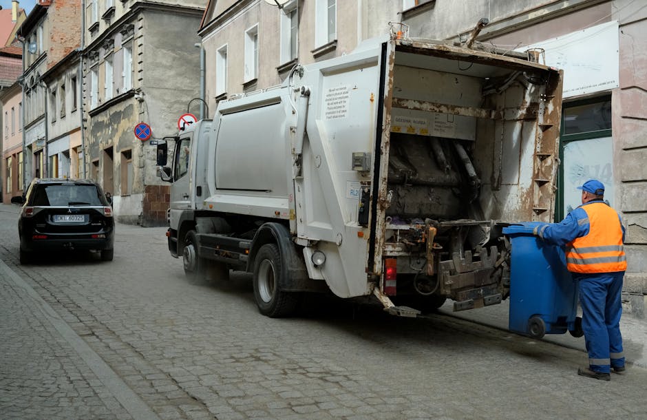 A waste collection truck, primarily white with some rusted metal components at the rear, is parked on a narrow cobblestone street next to a row of older, weathered residential buildings with beige and light gray facades. The truck's rear hatch is open, revealing an empty, enclosed compartment with visible rust, peeling paint, and mechanical parts used for compacting waste. A worker wearing a blue uniform, high-visibility orange vest, matching blue cap, and gloves is standing beside the truck, manoeuvring a blue wheeled bin towards the open rear. The street environment is urban and quiet, with minimal traffic; a small black car is parked further along the curb, and there are no other pedestrians visible. The scene is naturally lit by daylight, emphasizing the textures of the cobblestones, the worn building exteriors, and the metallic surfaces of the rubbish removal equipment. This setup demonstrates a typical independent collection or on-site waste disposal process managed by a waste management service like Rubbish Removal Harringay, reflecting an alternative to local authority rubbish collection.