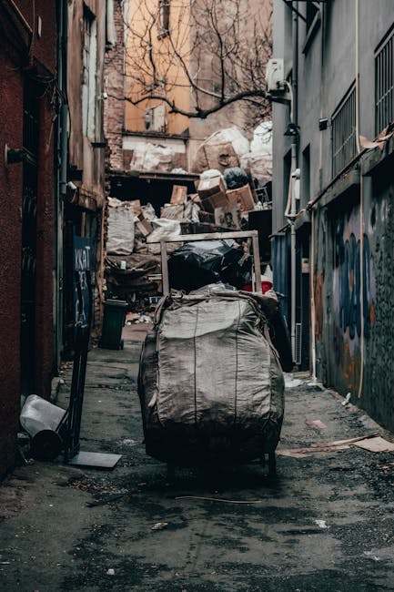 A narrow urban alleyway lined with graffiti-covered walls on the right and a metal fence on the left, leading to a piled accumulation of various types of rubbish and waste materials at the far end. In the foreground, a large, grey, weathered rubbish sack with visible creases and stains stands upright on the uneven pavement, with a metal shopping trolley partially covered in debris nearby. Behind the trolley, an assortment of cardboard boxes, plastic bags, and loose waste are stacked haphazardly, creating a cluttered scene typical of private waste disposal sites or on-site clearance efforts. The background features leafless trees and a mix of residential and commercial building facades, suggesting an inner-city environment. The lighting is subdued, casting soft shadows and highlighting the textures of trash, brickwork, and graffiti, emphasizing the scene’s nature as a snapshot of waste accumulation and the importance of rubbish removal services such as those offered by Rubbish Removal Harringay.