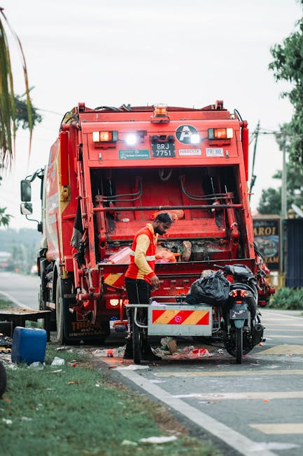 A worker in a yellow and red uniform is engaged in rubbish collection at the rear of a large red rubbish truck positioned on a roadside. The truck has a metallic, industrial finish with a red paint that appears slightly weathered, and it features various safety and identification labels, including a registration number 'BRJ 7751.' The worker is leaning forward, loading or inspecting waste into the open rear compartment, which is equipped with hydraulic mechanisms for compacting rubbish. On the ground nearby, there is a black plastic bag filled with waste, along with a few scattered debris and a blue container, suggesting on-site clearance or waste collection activity. The scene is set in a suburban area with a paved street, a grassy verge on the left, and a few trees visible in the background, along with power lines and a sign for a local business. The lighting indicates daytime with a cloudy sky, giving a neutral, natural atmosphere typical for street cleaning or refuse collection in a residential or commercial zone, aligning with independent rubbish removal services offered by Rubbish Removal Harringay.