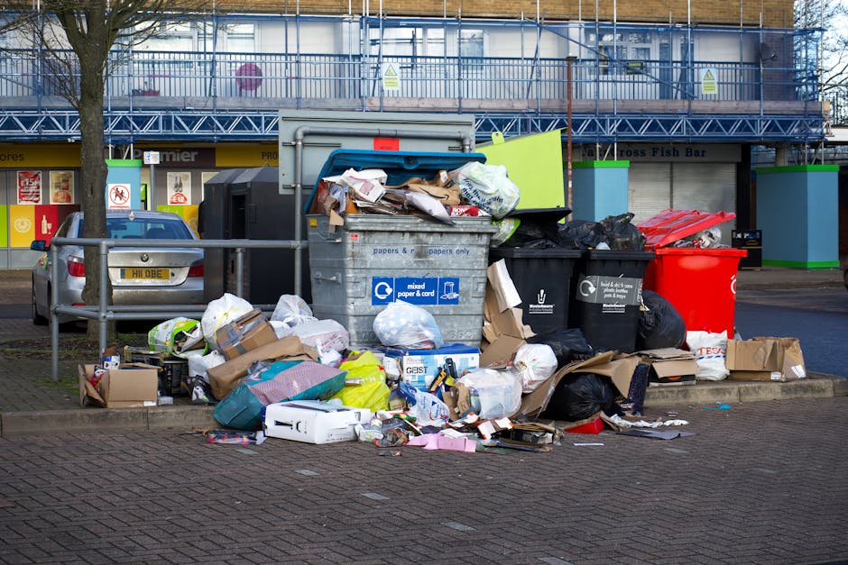 A worker in a yellow and red uniform is engaged in rubbish collection at the rear of a large red rubbish truck positioned on a roadside. The truck has a metallic, industrial finish with a red paint that appears slightly weathered, and it features various safety and identification labels, including a registration number 'BRJ 7751.' The worker is leaning forward, loading or inspecting waste into the open rear compartment, which is equipped with hydraulic mechanisms for compacting rubbish. On the ground nearby, there is a black plastic bag filled with waste, along with a few scattered debris and a blue container, suggesting on-site clearance or waste collection activity. The scene is set in a suburban area with a paved street, a grassy verge on the left, and a few trees visible in the background, along with power lines and a sign for a local business. The lighting indicates daytime with a cloudy sky, giving a neutral, natural atmosphere typical for street cleaning or refuse collection in a residential or commercial zone, aligning with independent rubbish removal services offered by Rubbish Removal Harringay.
